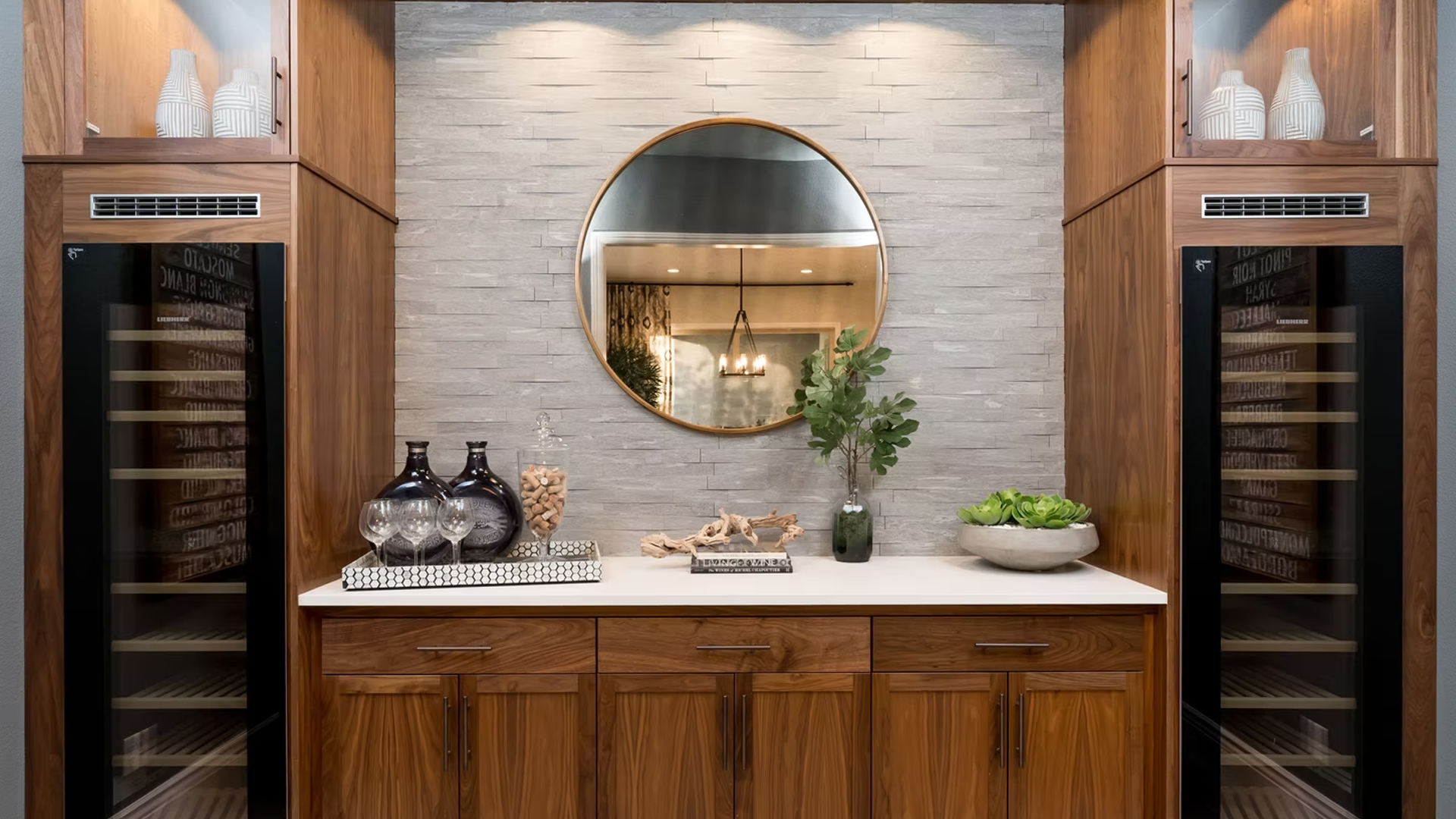 Built-in bar with wood cabinets, wine coolers, a stone wall, round mirror, and counter with decor.