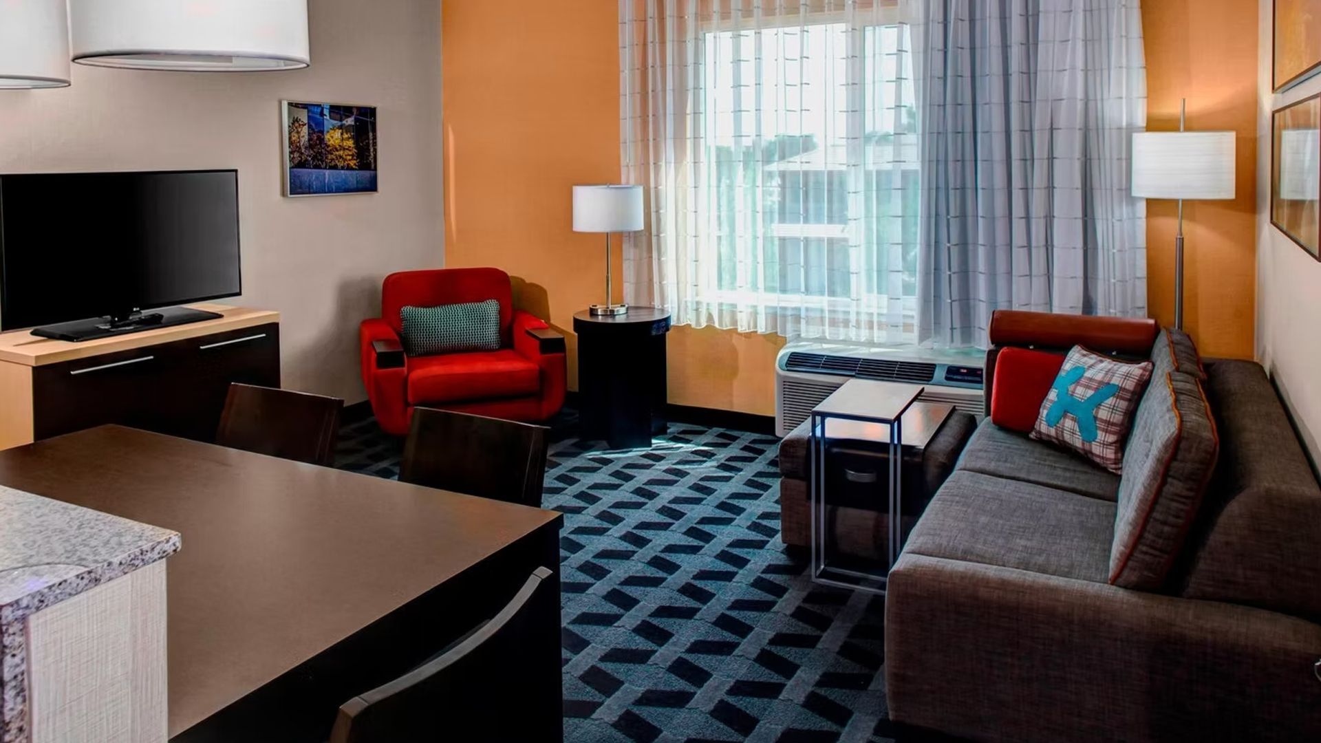 Hotel suite living area: TV, sofa, red chair, orange wall, patterned carpet, window, dining table.