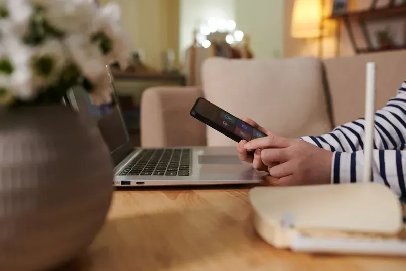 Person Holding Phone Near Laptop and Router on a Table With Flowers — Superior Wifi in Lismore, NSW