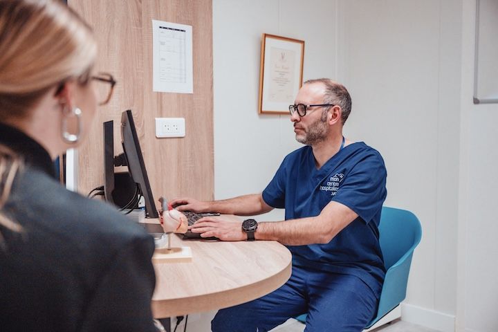 Un médecin est assis à une table et parle à un patient tout en utilisant un ordinateur.