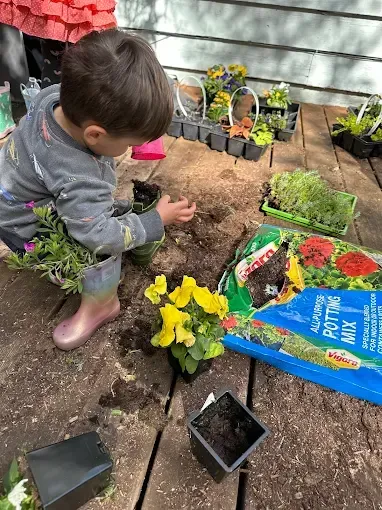 Young child planting flowers on a wooden deck with gardening supplies.