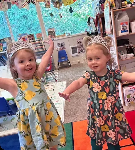 Two young girls in dresses and tiaras, smiling and waving indoors, possibly a classroom.