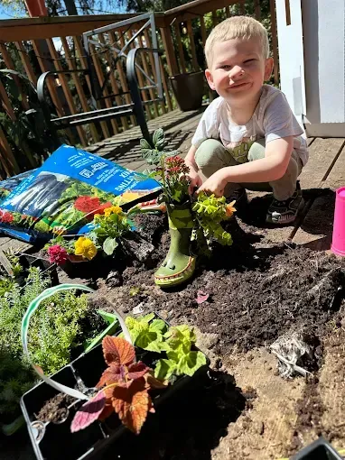 Young child gardening, kneeling and smiling, planting flowers in a green boot; dirt, plants, and soil bag.