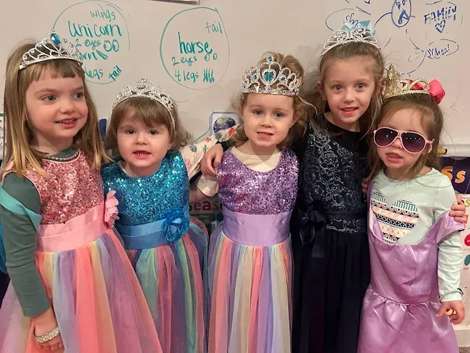 Five young girls in princess dresses and tiaras posing together.