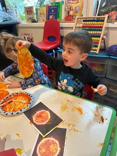 Child excitedly holds up orange-colored art, making a sun. Two other children work on art, in a classroom.