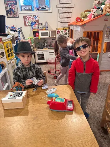 Three children playing shop in a playroom. One counts toy money, another wears sunglasses, and a third plays near a toy kitchen.