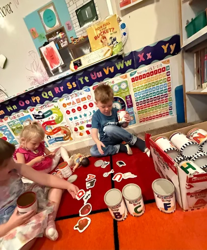 Children playing with cans and alphabet letters on a red rug in a classroom.