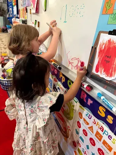 Two young girls writing on a whiteboard. One has blonde hair, the other black. They are in a classroom.