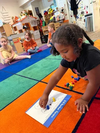 Young girl placing blocks on a worksheet during a number review activity in a classroom. Other children nearby.