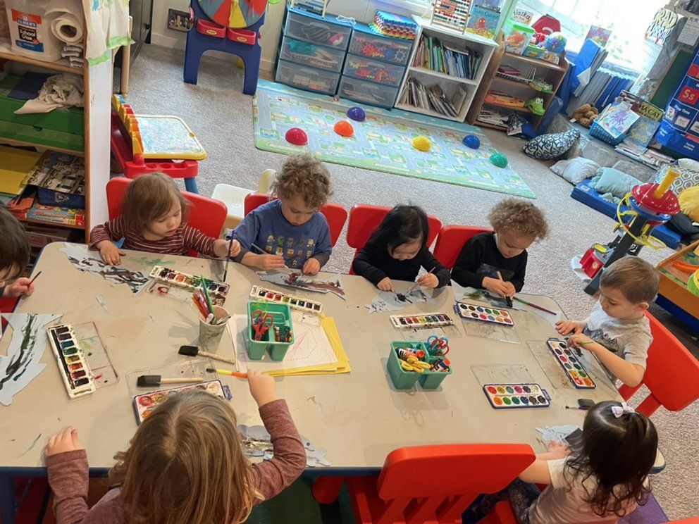 Children painting at a table in a classroom; red chairs, watercolors, and a colorful rug are visible.