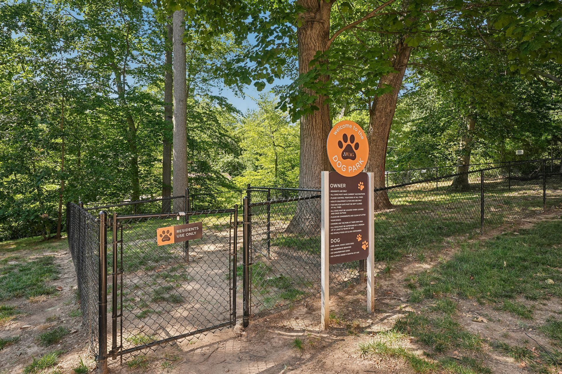 Dog park entrance gate with orange sign, surrounded by trees and greenery