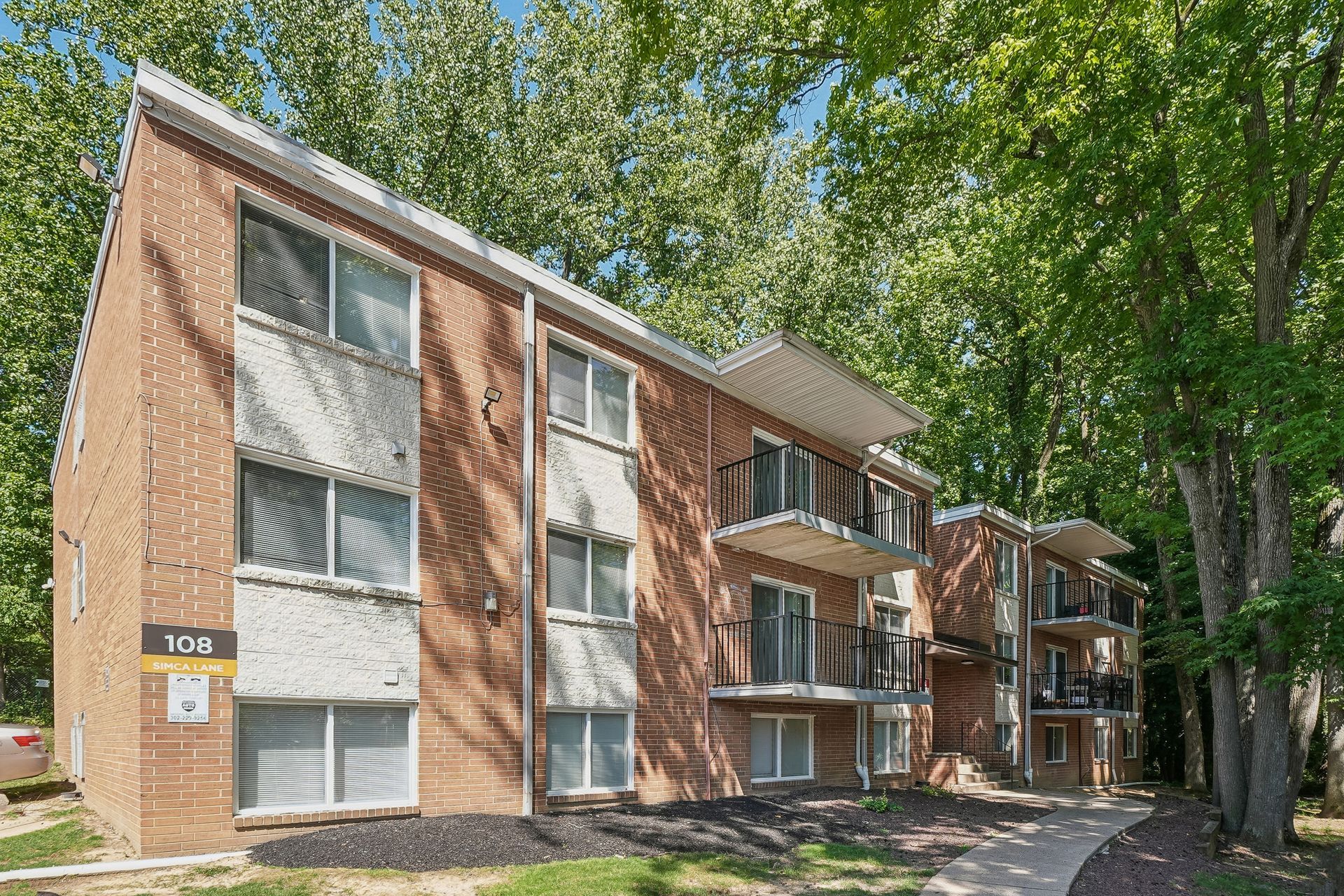 Brick apartment building with balconies and trees along a shaded walkway