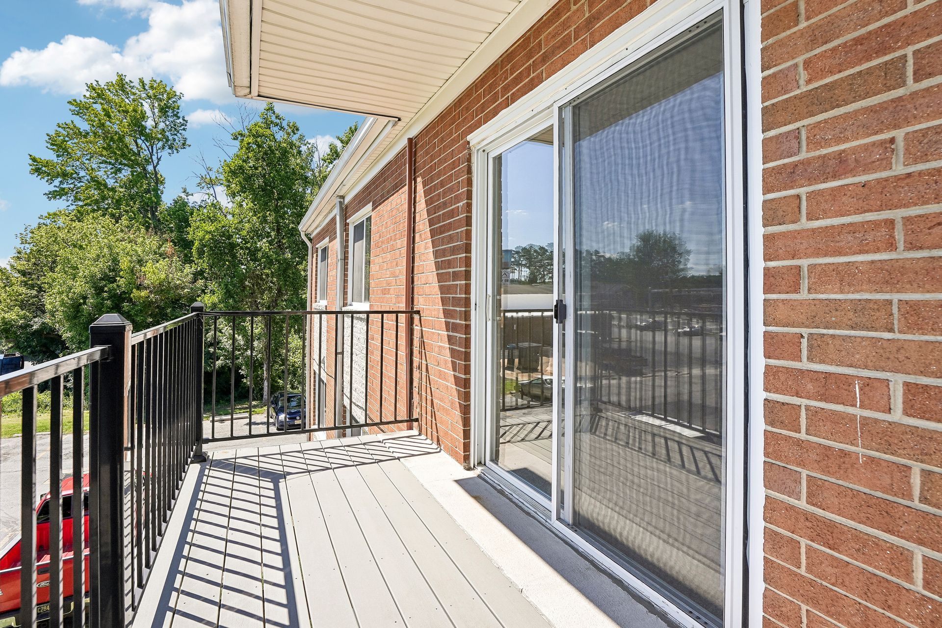Brick apartment balcony with black railing, white deck, and sliding glass doors in sunlight