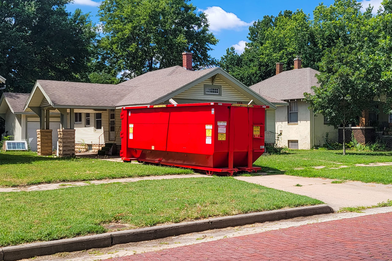 Red dumpster sits on a driveway in front of a house, with green grass and trees in the background.