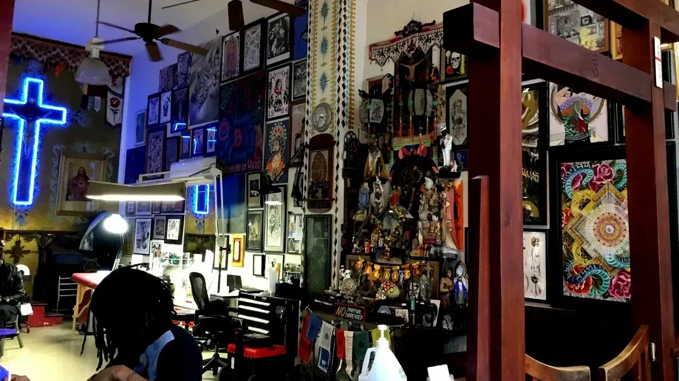 A man is sitting in a tattoo parlor with a neon cross on the wall.