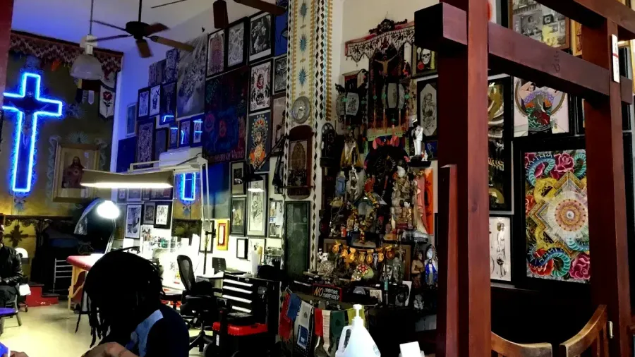 A man is sitting in a tattoo parlor with a neon cross on the wall.