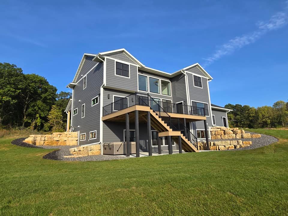 A large house with a deck and stairs is sitting on top of a lush green field.