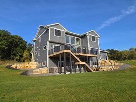 A large house with a deck and stairs is sitting on top of a lush green field.