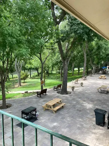 Outdoor patio with picnic tables, benches, and a grill under trees near a river.
