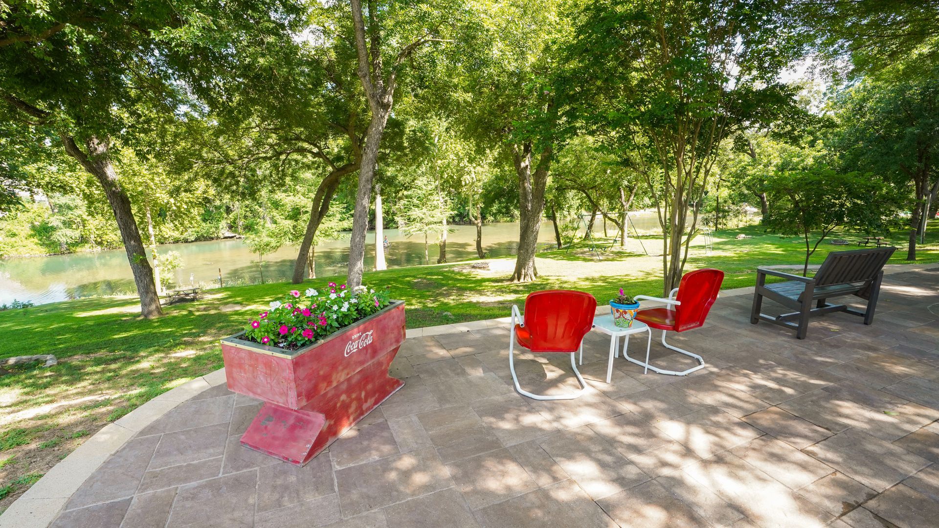 A patio with red chairs, a flower box, and a view of a river under shady trees.