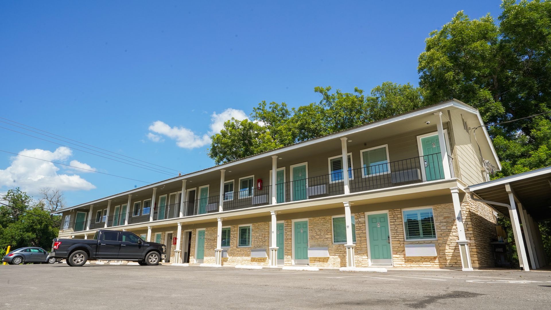 Two-story motel with light brown brick facade, teal doors, and blue sky. A truck is parked out front.