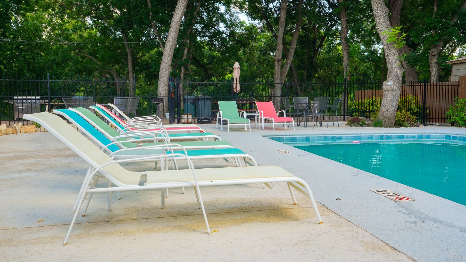Poolside with lounge chairs. Some are teal, green and pink, alongside a pool with trees in the background.