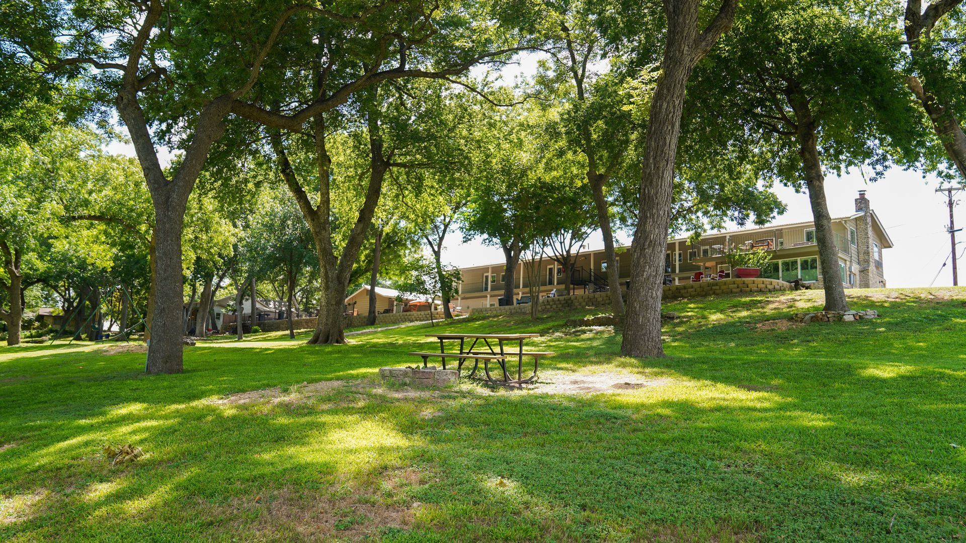 Picnic table under trees in a park, with a building visible in the background. Green grass and foliage.