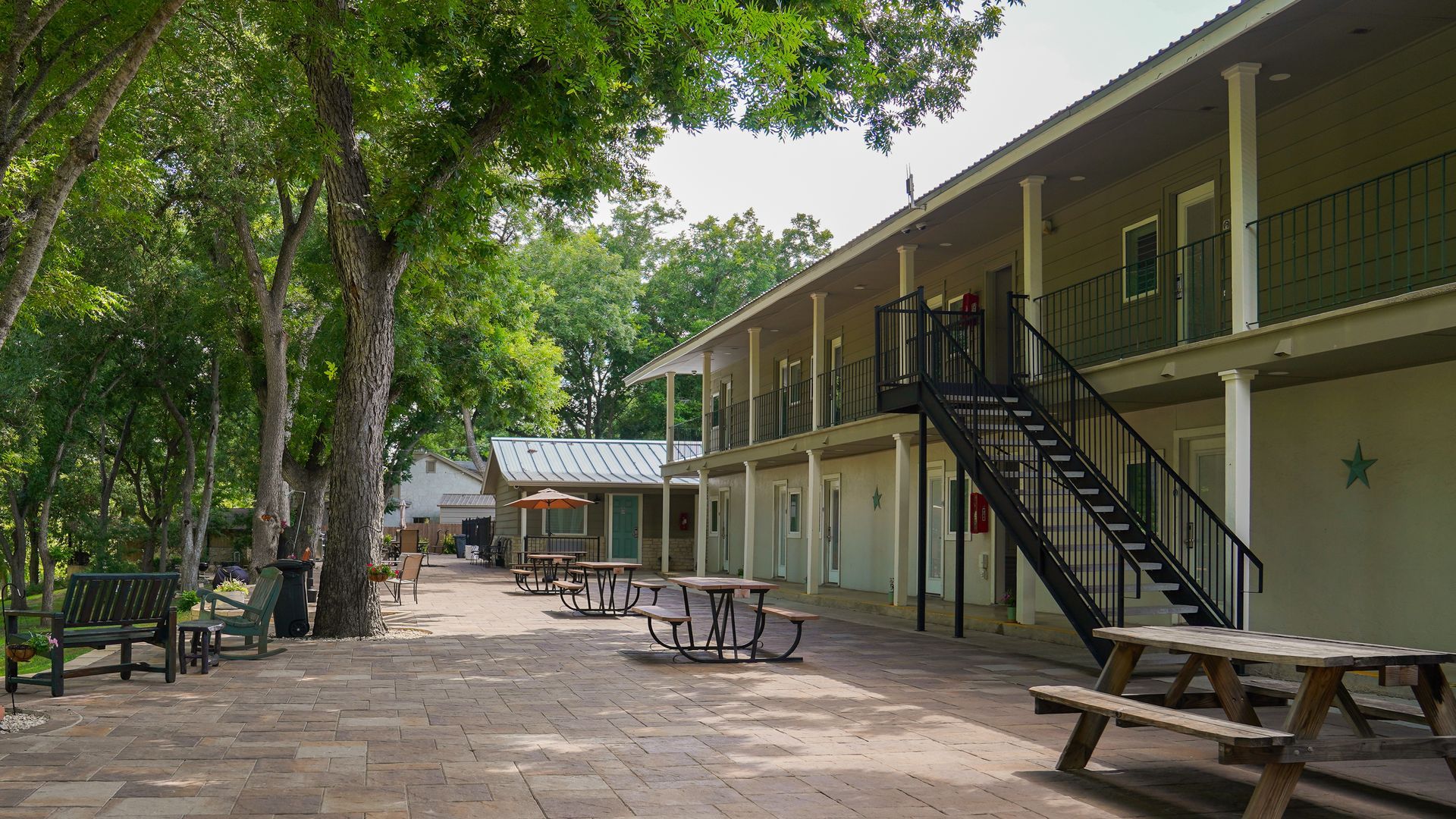 Patio with tables and chairs in front of a two-story building with black staircase and trees.