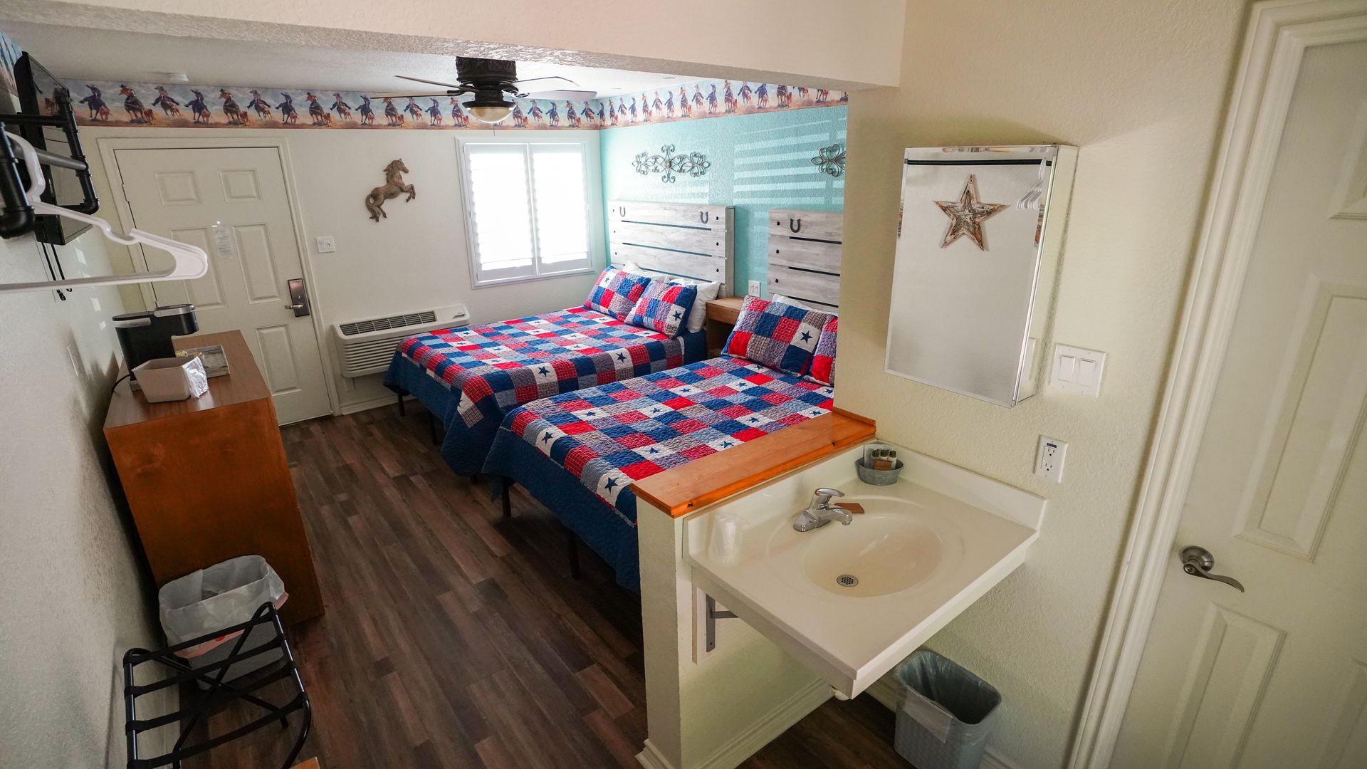 Hotel room with two twin beds, a sink, and a door. The room has a western theme with a window and a television.
