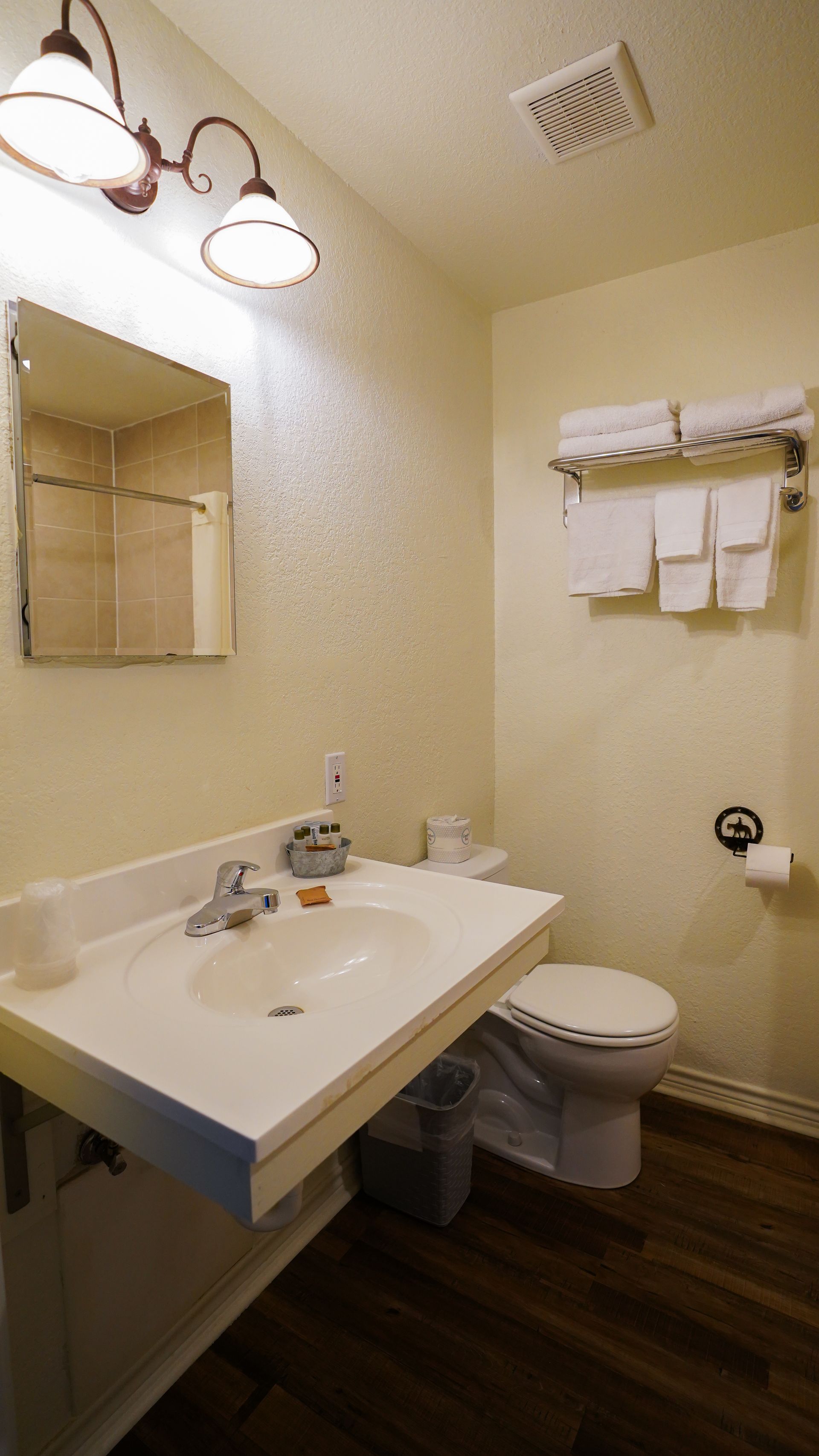 Bathroom with white sink, toilet, towels, and mirror; speckled wallpaper and wood-look floor.