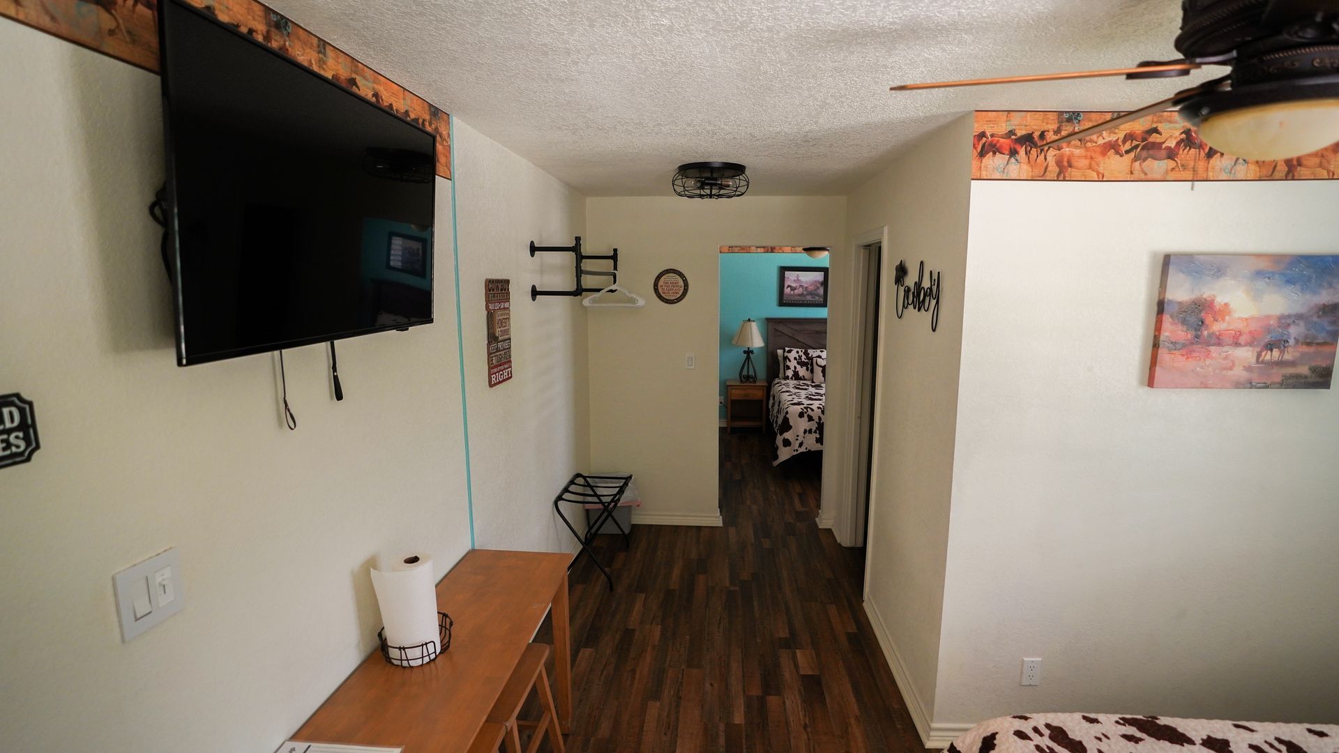 Narrow hallway with a TV, shelf, luggage rack, and a door to a bedroom with a bed.