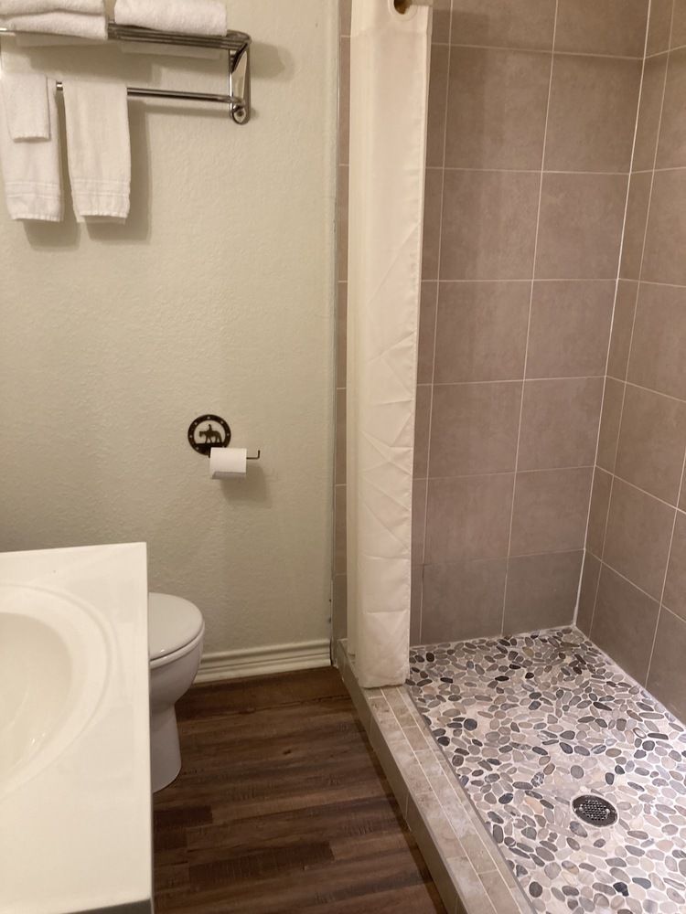 Bathroom with toilet, shower, sink, and towels. Brown wood floor, grey tile shower, and white fixtures.