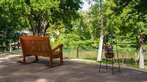Wooden rocking bench overlooking a river, with a hanging flower basket on a metal stand.