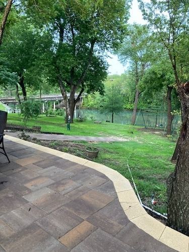 Patio overlooking a grassy yard, trees, and a river. Brown and gray pavers with green grass.