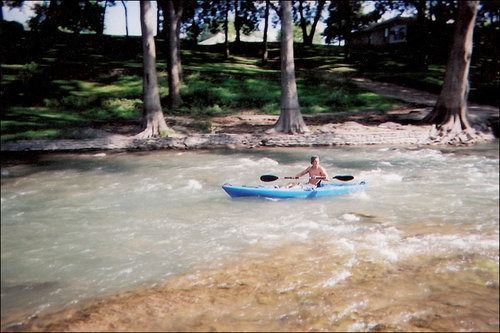 Person kayaking in a blue kayak on a river; trees and green foliage in the background.