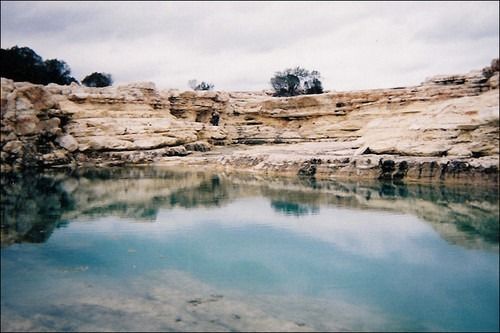 A tranquil, turquoise water-filled quarry reflects the tan, layered rock walls and cloudy sky.