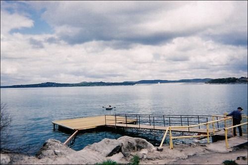 Wooden pier extends into a large lake under a cloudy sky. Person on the pier. Boat on water.