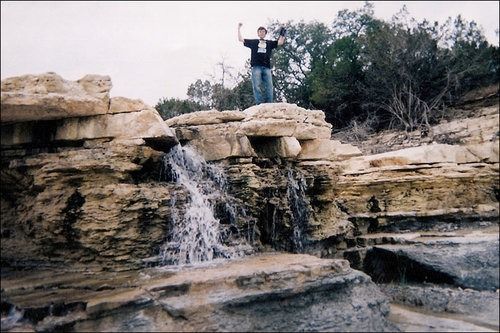 Person with arms raised on rocky ledge above small waterfall.