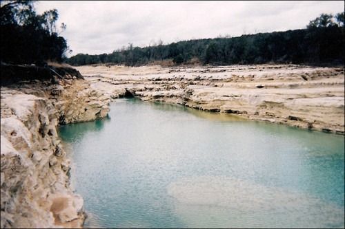 River with turquoise water and light-colored banks, surrounded by trees under a cloudy sky.