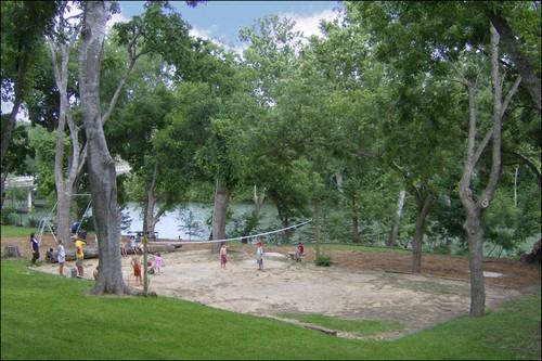 Children playing volleyball in a sandy area surrounded by trees and a grassy area.