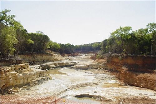 Dry riverbed flanked by eroding banks, trees overhead. Sunny day.