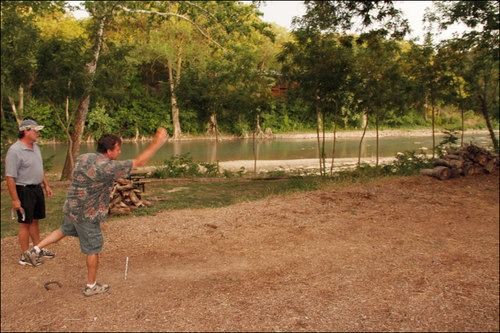 Two men playing horseshoes near a river. One throws, the other watches, trees in the background.
