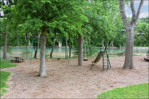 Playground with swings and slide by a river, surrounded by trees.