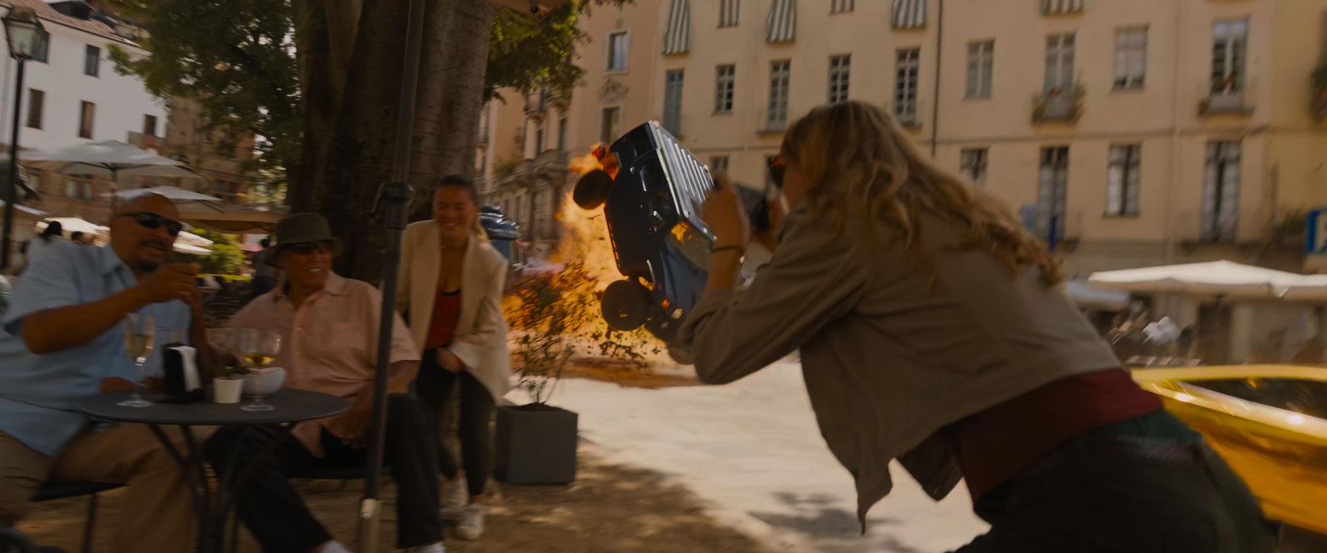 Fast X Fast and Furious 10 action scene in Rome with a massive explosion and a car flipping in the background, while a group of tourists poses for a photo in the foreground, seen from the viewpoint of camera operator Ferdi Fischer.