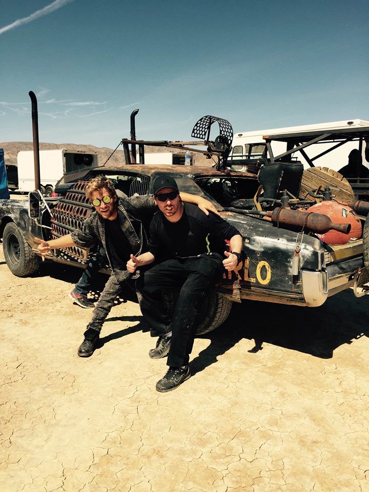 Ferdi Fischer and another stunt performer standing side by side in the Nevada desert on a dry lakebed, blue sky above and several heavily modified Mad Max style vehicles parked behind them on the sand.