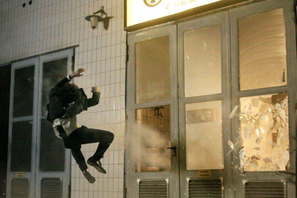 Stunt performer Ferdi Fischer midair in front of a large garage door with windows on a Black Forest movie set, being yanked backwards by a ratchet pullback at the exact moment the explosion blast shatters the glass panels behind him.