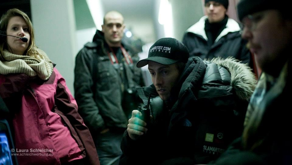Action director Ferdi Fischer standing at Video Village on a film set in Germany, holding a radio and watching the monitor closely with several crew members gathered around him as they review a fight scene in progress.