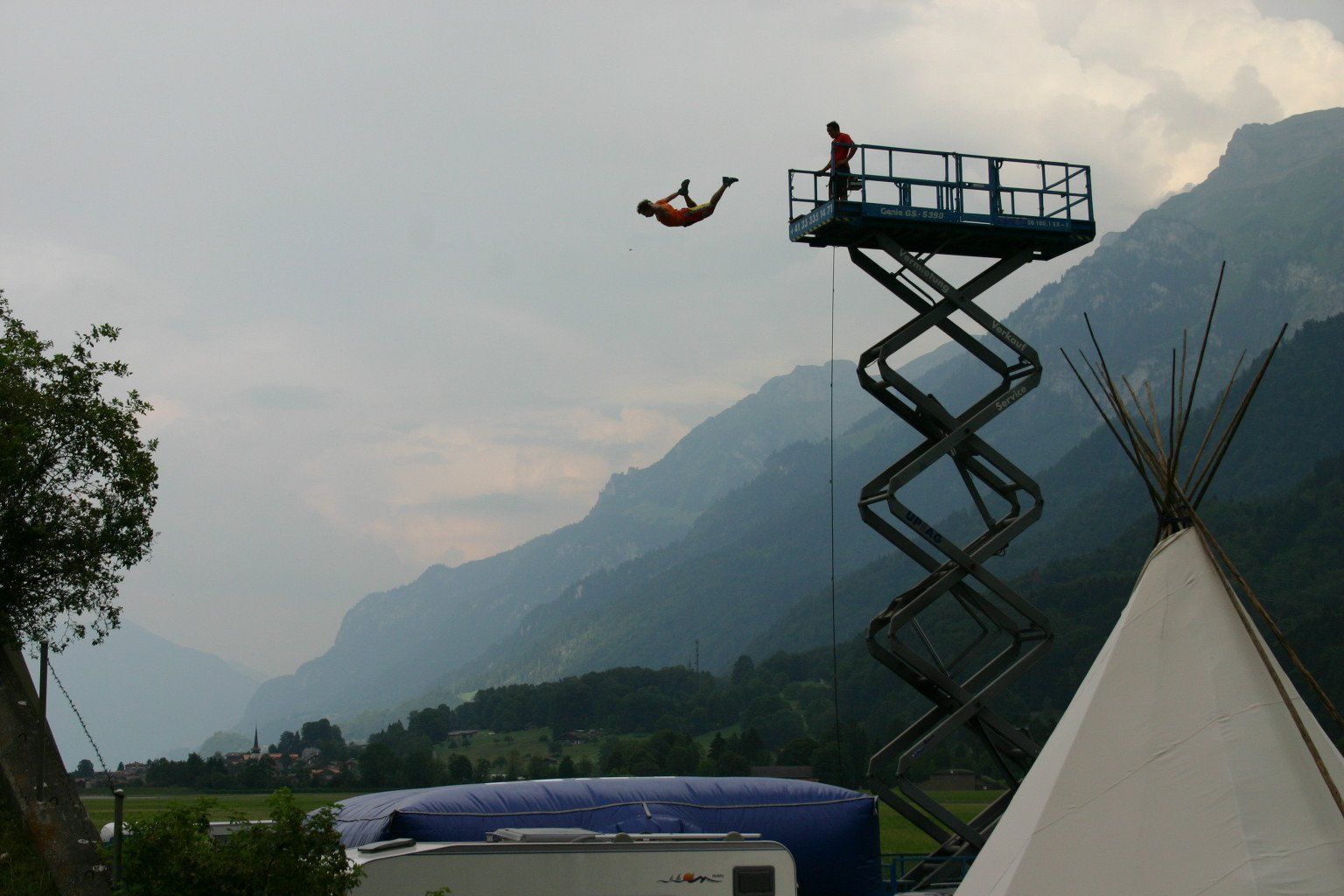 Action director and stunt performer Ferdi Fischer jumping from a scissor lift roughly 10 to 15 meters above the ground into an airbag during a Stunt Hero TV show shoot in Interlaken, with a scenic backdrop of Swiss mountains and valleys behind him.
