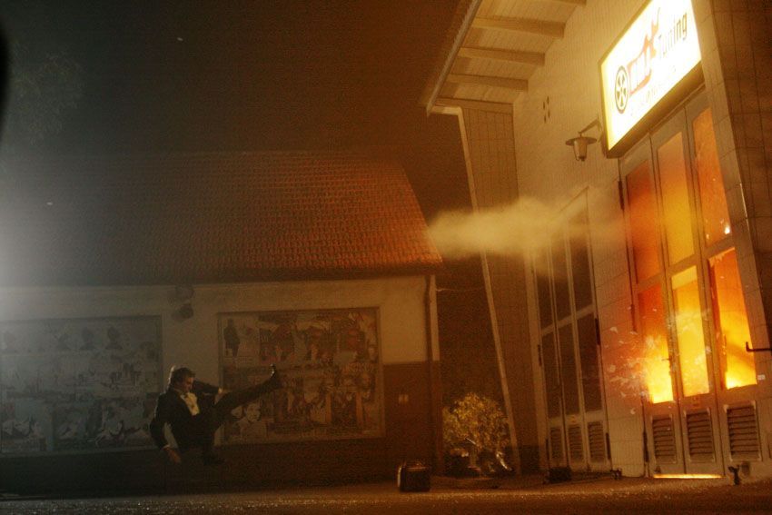 Side angle stunt shot of Ferdi Fischer suspended in the air in front of a garage door on a Black Forest movie set, a split second after the blast as bright orange flames and debris burst through the windows behind him, moments before he is fully engulfed by the explosion.