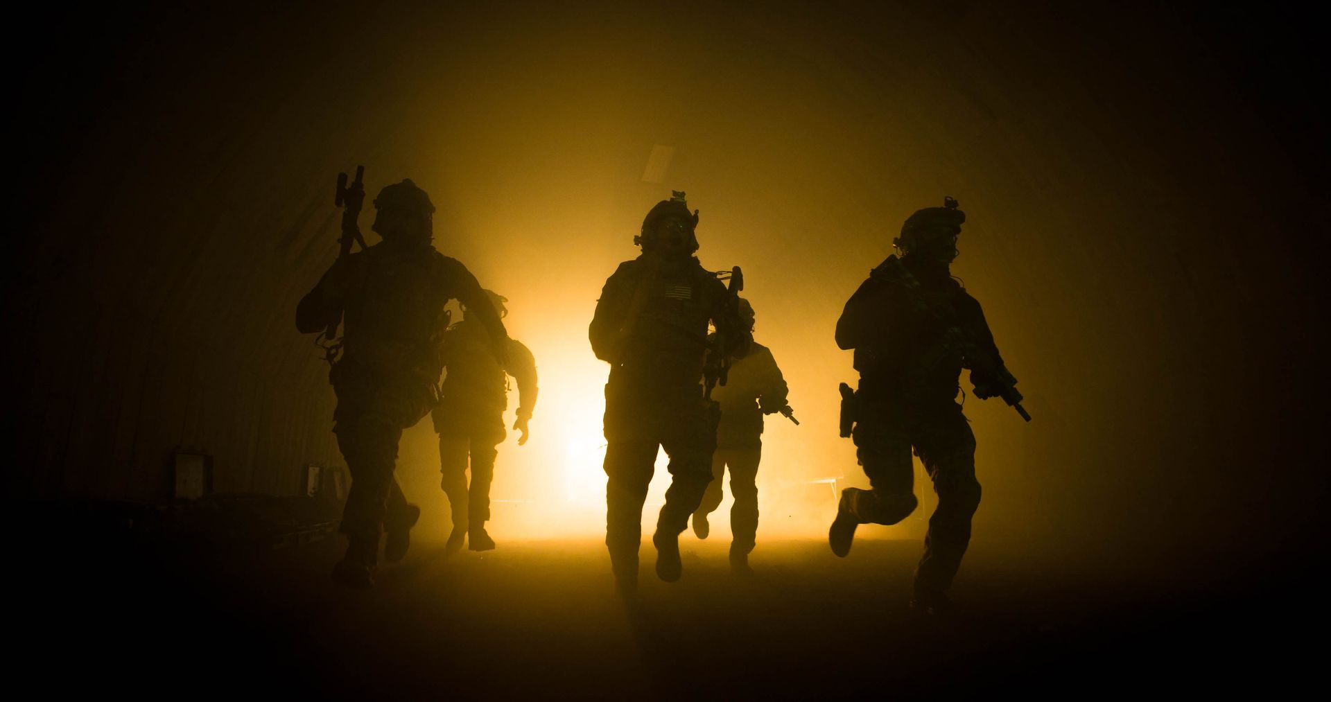 Image of a group of military operators in tactical gear advancing cautiously in a combat zone, with their weapons drawn and ready.